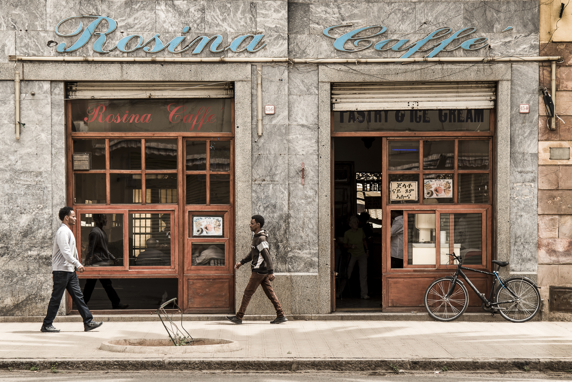 LO STRANO CASO DI REIRATE. Due persone camminano davanti al "Rosina Caffè" in un’ambientazione che ricorda l’Italia di qualche decennio fa. Questa è Asmara in Eritrea, ex-colonia italiana, dichiarata Patrimonio dell’Unesco nel 2017. ©Cristiano Zingale